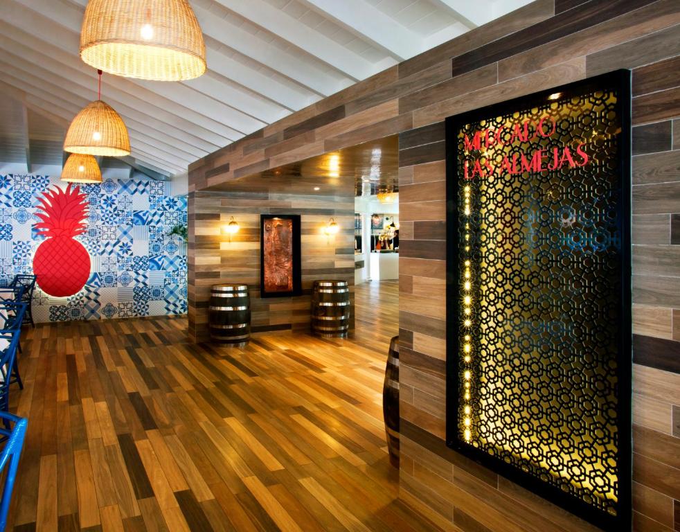 Moderns restorāns ar koka grīdu un dekoratīvu sienu, kurā redzama sarkana ananāsu ilustrācija. Interjera dizainā ir izmantoti dabiski materiāli un eleganti apgaismojumi.