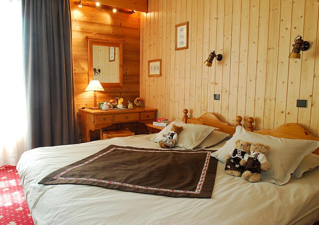 Mūsdienīgs guļamistabas interjers ar koka sienām, ērtu gultu un mīkstiem spilveniem. Uz gultas novietoti divi mīkstie lāči un blakus esošs koka galds ar lampu un dekoratīviem priekšmetiem.