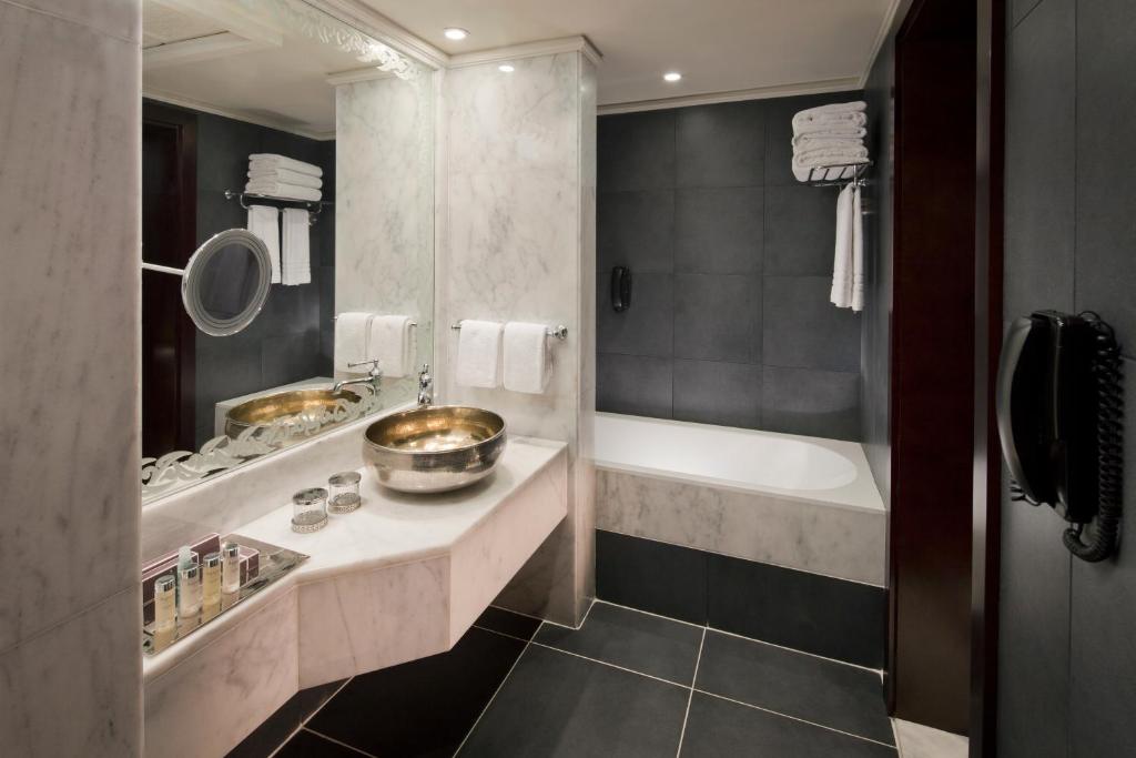 Mūsdienīgs vannas istabas interjers ar marmora izlietni, sudraba trauku un melnām flīzēm. Uz galda novietoti kosmētikas līdzekļi un dvieļi, radot elegantu un funkcionālu atmosfēru.