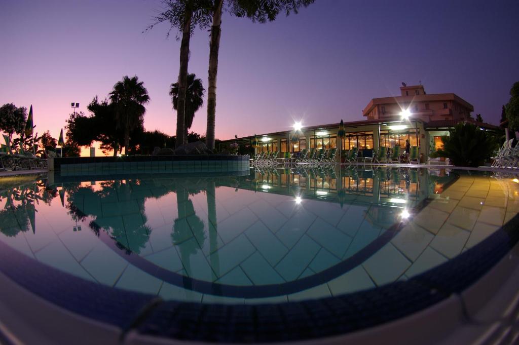 Peldbaseins ar miera atmosfēru, apgaismots saulrieta gaismā, ar palmām un viesnīcu fona. Ideāla vieta relaksācijai un atpūtai.