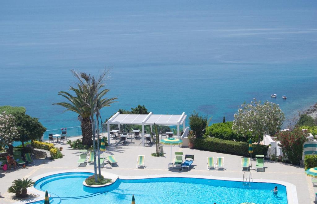 Viesnīcas baseins ar saulessargiem un pludmales krēsliem, skats uz zilo jūru un krasta līniju. Apkārtne ir zaļa ar palmām un ziedošām augiem.