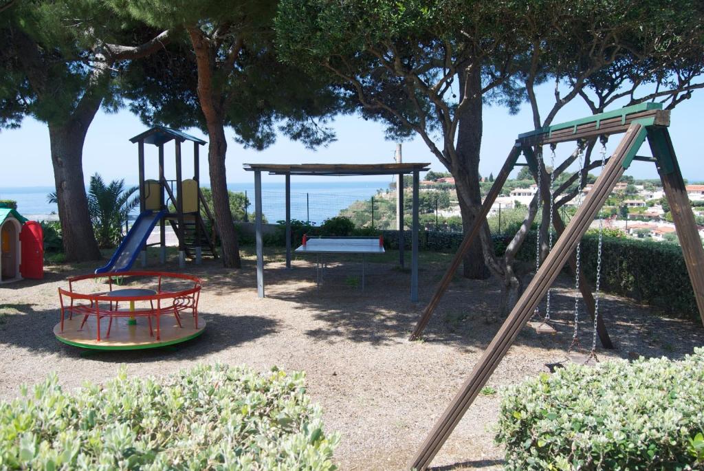 Bērnu rotaļu laukums ar šūpolēm, slide un griezošo platformu, atrodas saulainā vietā ar skatu uz jūru. Apkārt aug koki un krūmi, radot patīkamu atmosfēru.