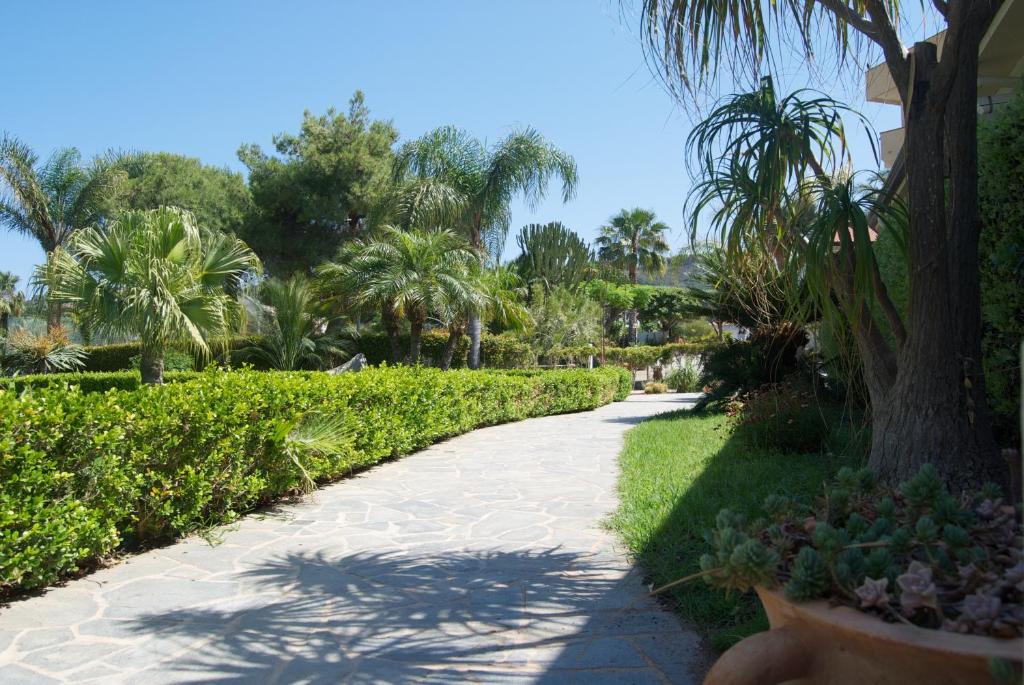 Skaists dārza ceļš, ko ieskauj zaļi augi un palmas, kas rada mierīgu un relaksējošu atmosfēru. Dabas skaistums un saulains laiks padara šo vietu ideālu pastaigām.