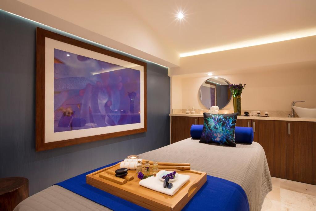 Spa telpa ar masāžas galdu, dekoratīvu spilvenu un relaksējošu atmosfēru. Sienā ir krāsains mākslas darbs, kas piešķir telpai mājīgu noskaņu.