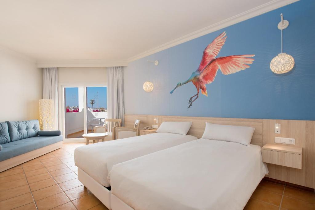 Mūsdienīgs viesnīcas numurs ar divām gultām, gaišām krāsām un krāšņu putna attēlu uz sienas. Telpā ir moderna mēbele, dabiskais apgaismojums un piekļuve balkonam ar skatu uz dārzu.