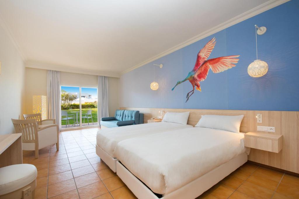 Mūsdienīga guļamistaba ar diviem gultām, spilgti zilu sienu un krāsainu putnu attēlu. Lielas logu durvis ved uz dārzu, radot gaišu un patīkamu atmosfēru.