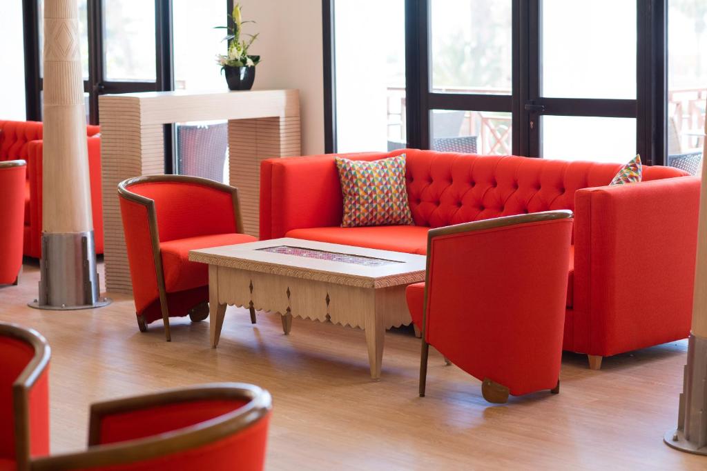 Mūsdienīgs viesnīcas interjers ar spilgtām sarkanu krāsu mēbelēm, tostarp dīvānu un krēsliem, apvienots ar elegantu koka galdu. Interjers rada siltu un viesmīlīgu atmosfēru.