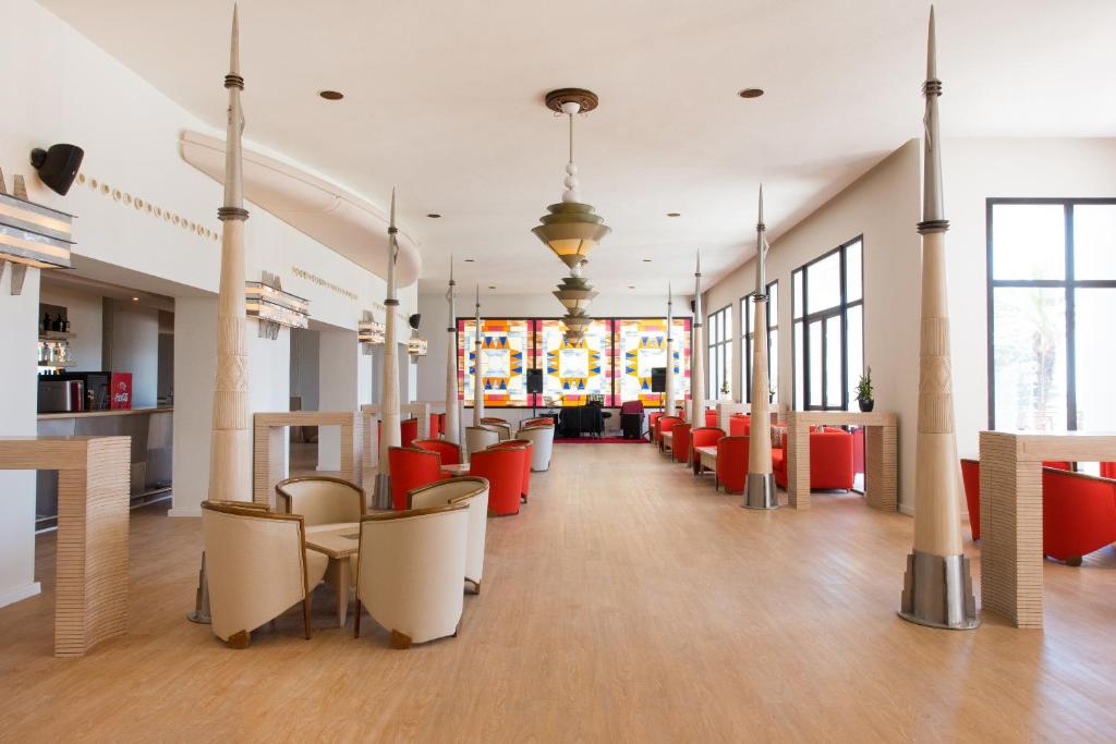 Mūsdienīga un eleganta interjera telpa ar sarkaniem krēsliem, koka grīdu un interesantiem gaismas ķermeņiem. Lielie logi nodrošina dabisko apgaismojumu, radot patīkamu atmosfēru.