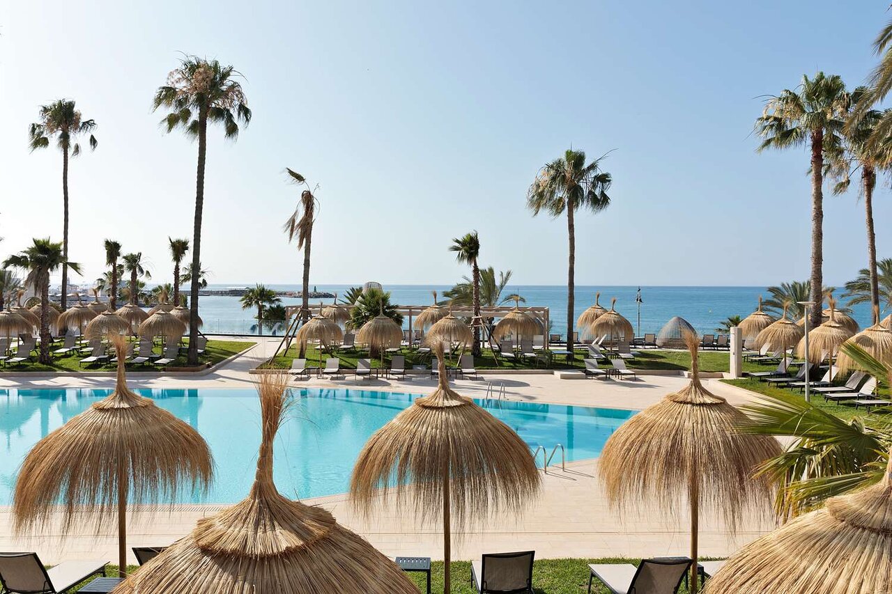Tropu pludmales skats ar palmām, peldbaseinu un sauļošanās vietām, izveidojot relaksējošu atmosfēru. Jūras horizonts papildina idillisko ainavu. panoramic view with beach and pools