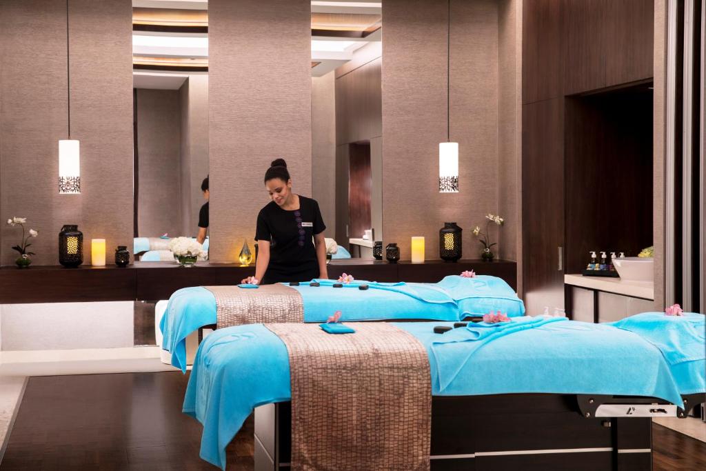 Spa telpa ar masāžas galdiem un relaksējošu atmosfēru, kur masāžas terapeits sagatavo procedūras. Interjerā ir mīksti toņi un dekoratīvi elementi, kas veicina mieru.