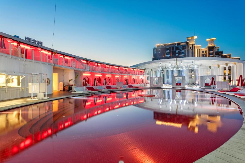Mūsdienīgs baseins ar sarkanu apgaismojumu un stilīgām atpūtas zonām, kas atspoguļojas ūdenī. Apkārt ir eleganti sēdvietas un moderna arhitektūra.