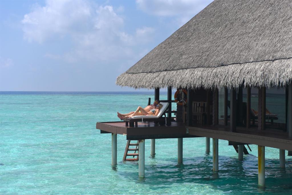 Atpūtas vieta pie jūras ar jumtu no salmiem, kur cilvēki atpūšas uz terases virs kristāldzidriem ūdeņiem. Ideāla vide relaksācijai un brīvdienām.