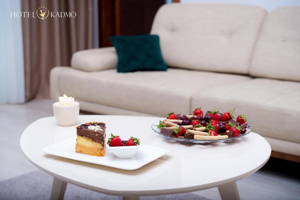 Saldā kūka un augļu plate ar zemenēm un vīnogām uz galda pie dīvāna. Interjers ir mūsdienīgs un mājīgs, radot patīkamu atmosfēru.