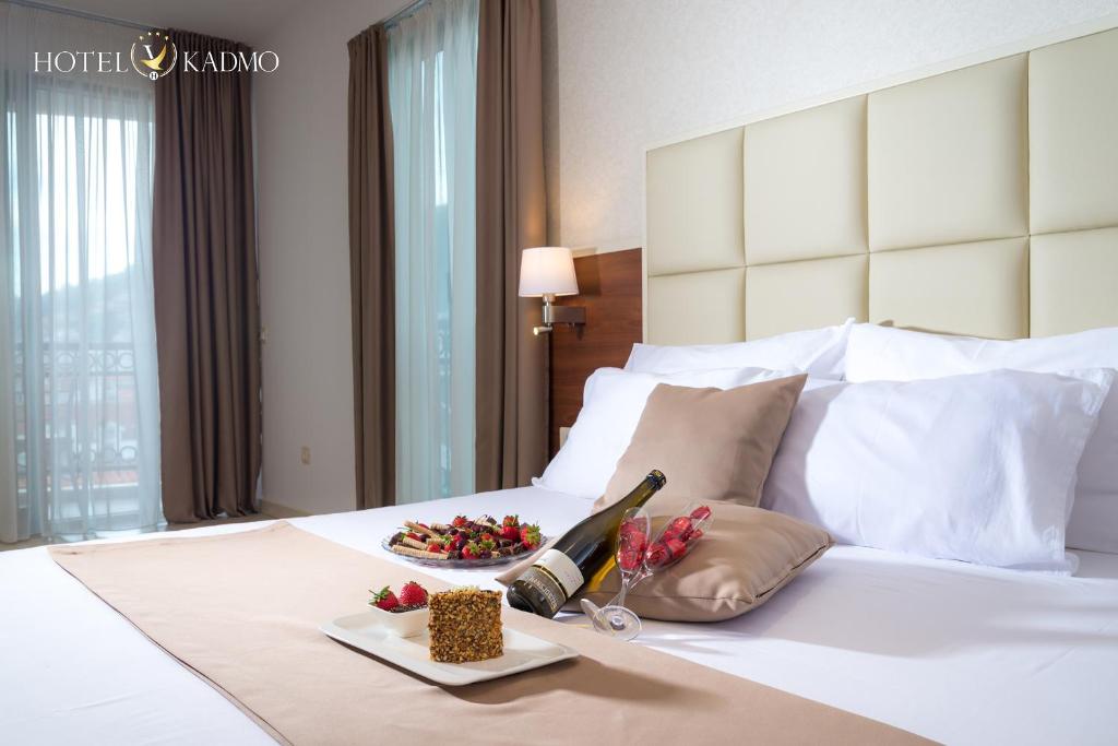 Elegants viesnīcas numurs ar gultu, apklātu ar baltām gultasveļām un dekoratīvām spilveniem. Uz gultas redzama šampanieša pudele un desertu plates ar augļiem. Aiz logiem plūst dabiska gaisma, radot mājīgu atmosfēru.