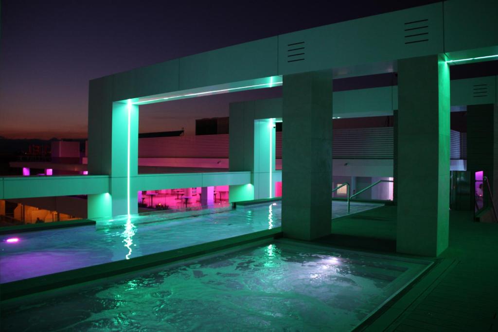 Mūsdienīgas arhitektūras peldbaseins ar krāsainu apgaismojumu, kas izceļ stilīgas līnijas un ūdens virsmu. Vakara gaisotnē redzamās zaļās un rozā gaismas piešķir unikālu atmosfēru.