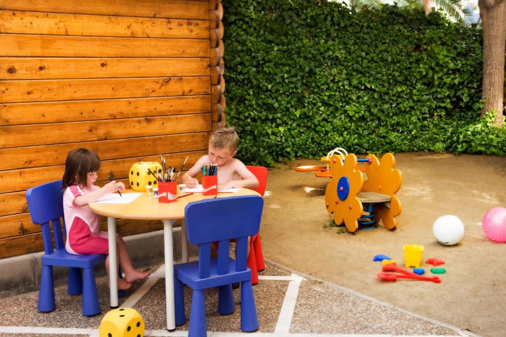 Bērni radoši darbojas pie galda, zīmējot un krāsojot. Apkārtnē ir rotaļlietas un zaļš fons, kas rada priecīgu un radošu atmosfēru.