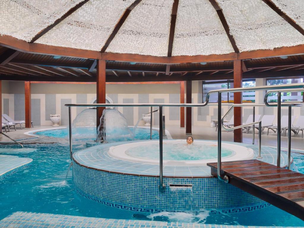 Relaksējoša spa telpa ar baseiniem un burbuļvannām, kurā apmeklētājs baudīšanas brīdis ūdenī. Dabas gaisma plūst caur jumtu, radot mierīgu atmosfēru.