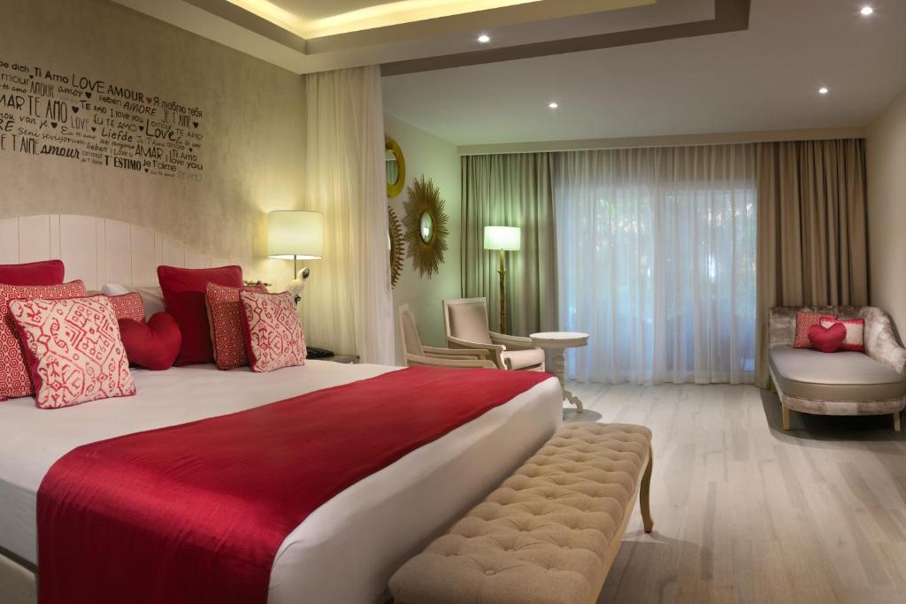 Mūsdienīgs guļamistabas interjers ar sarkaniem akcentiem un romantisku atmosfēru. Telpā ir plaša gulta ar dekoratīvām spilvenām un mīksta dīvāna stūra zonas.