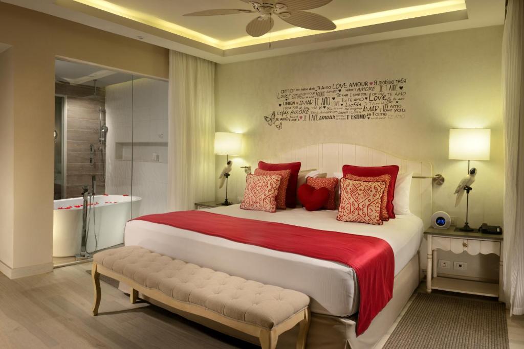 Romantisks viesnīcas numurs ar sarkanu gultas pārklāju un dekoratīvām spilveniem, grezns vannas istabas stūris ar ziedlapiņām un modernu apgaismojumu.