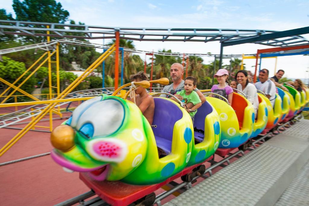 Krāsaina bērnu atrakcija ar cilvēkiem, kas priecīgi brauc uz kalniņiem, kas izskatās kā ķirzaka. Fonā redzams parks ar zaļiem kokiem.