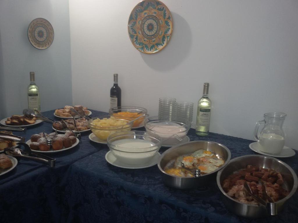 Bagātīga brokastu galda kompozīcija ar dažādiem ēdieniem, tostarp maizēm, augļiem, pienu un vīnu. Galda noformējums ar zilu galdautu un dekoratīvām šķīvju sienām.