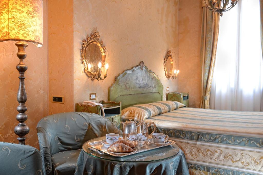 Eleganta viesnīcas numuriņa interjers ar gultu, krēsliem un galdu, uz kura ir pasniegti saldumi un dzēriens. Siltas krāsas un stilīgi apgaismojumi rada mājīgu atmosfēru.