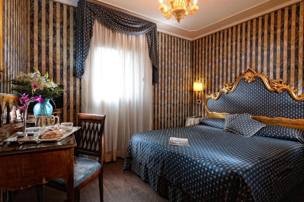 Eleganta viesnīcas istaba ar zilu un zeltītu apdari, dekoratīvu gultu un gaisīgu logu. Uz galda ir pasniegti brokastu našķi un ziedi, radot mājīgu atmosfēru.