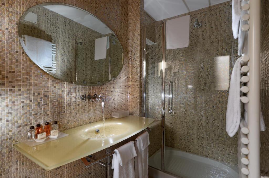 Mūsdienīgs vannas istabas interjers ar mozaīkas flīzēm, ovālu spoguli un dušas kabīni. Uz galda ir dāvanas un higiēnas līdzekļi.