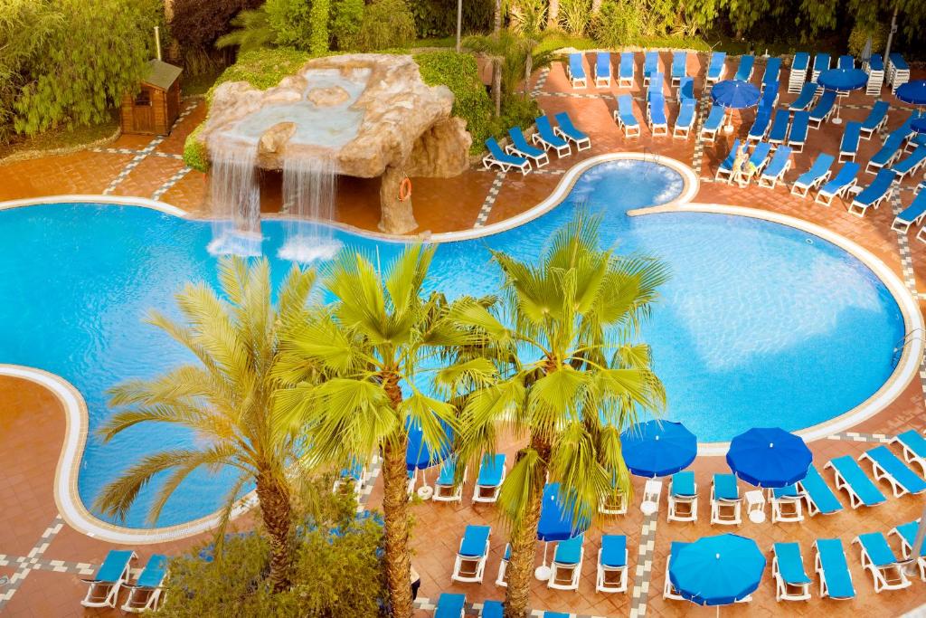 Peldbaseins ar ūdenskritumu un palmām, apkārtējā teritorijā redzami zili sauļošanās krēsli un lietussargi, radot relaksējošu atpūtas atmosfēru.