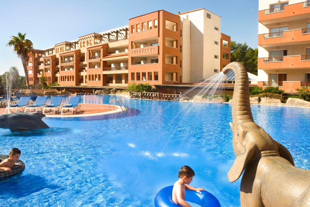 Baseins ar zilo ūdeni un bērniem, kas spēlējas, ar zilo gredzenu. Fons ar viesnīcas ēku un skulptūrām, piemēram, zilo delfīnu un zilo ziloņu.