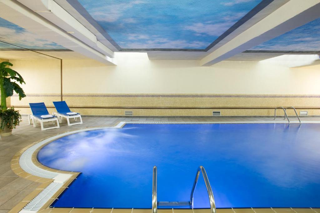 Iekštelpu peldbaseins ar zilu ūdeni un debesu motīvu griestiem, apkārtējo telpu papildina divas zāles ar baltiem krēsliem un zaļu augu.