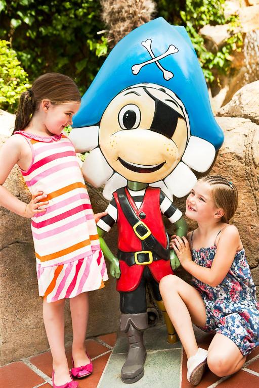 Divas meitenes pozē blakus krāsainai pirātu figūrai ar zilu cepuri. Viens no bērniem ir tērpies svītrainā kleitā, bet otrs valkā ziedu raksta kleitu.