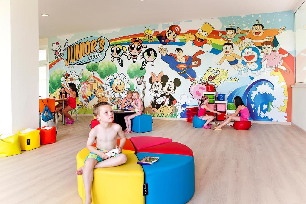 Bērnu klubs ar spilgtu sienu gleznojumu, kurā attēloti pazīstami multfilmu varoņi, un bērni spēlējas uz krāsainām mīkstajām mēbelēm. Telpa ir gaiša un draudzīga, radot priecīgu atmosfēru.