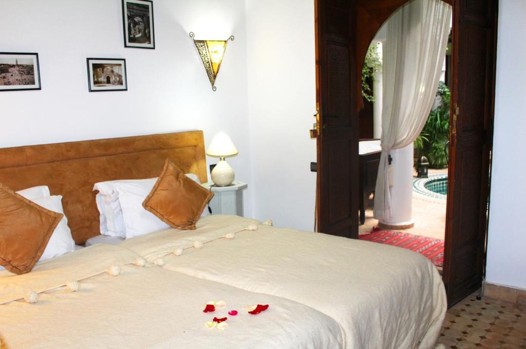 Mūsdienīga guļamistaba ar divguļamo gultu, dekoratīvām spilveniem un ērtu apgaismojumu, skatoties uz dārzu caur koka durvīm. Uz gultas ir rožu ziedlapiņas, pie sienas redzamas melnbaltas fotogrāfijas.