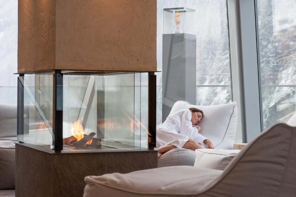 Relaksējoša ainava ar modernu kamīnu un ērtām sēdvietām, kur sieviete atpūšas baltā halātā, baudot mierīgu atmosfēru. Aiz loga redzama sniegota ainava.