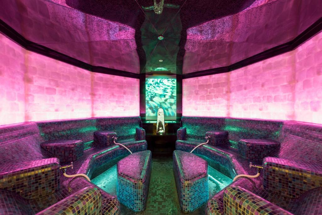 Mūsdienīga spa telpa ar krāsainām mozaīkas sienām un relaksējošām sēdvietām. Telpā ir maigs rozā un zaļš apgaismojums, radot mierīgu atmosfēru.