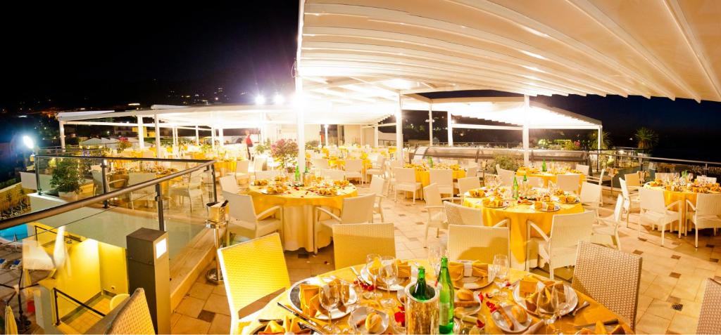 Eleganta restorāna terase ar izsmalcināti klātiem galdiem un zeltainām galdautām, kas izgaismota naktī. Apkārt ir zaļumi un baseins, radot patīkamu atmosfēru.