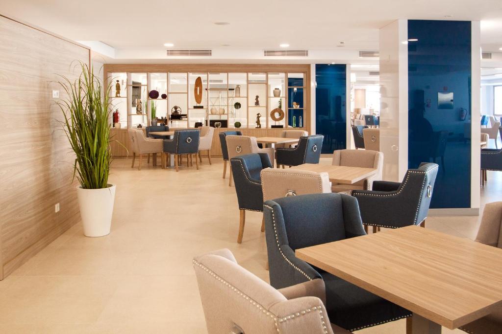 Moderns restorāns ar gaišām krāsām un elegantiem galdiem, kurā redzami ērti krēsli un dekoratīvi elementi. Rūpīgi izstrādāta interjera dizaina telpa.