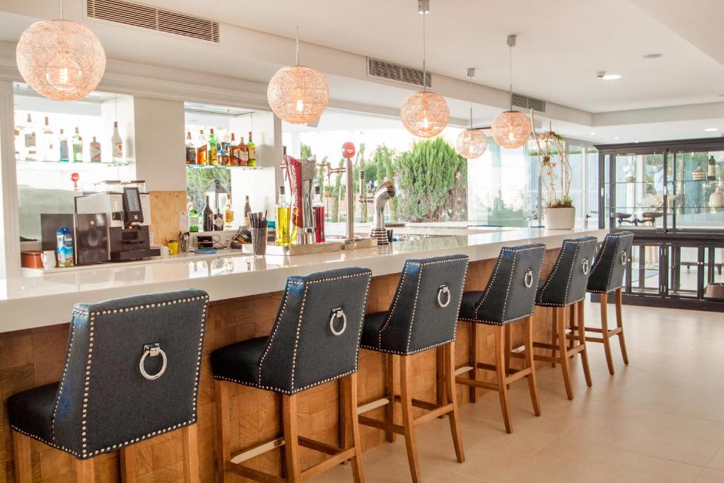 Mūsdienīgs bārs ar stilīgām melnām krēslām un mūsdienīgu apgaismojumu, kurā redzama dzērienu plaukti un eleganta baltā bāra virsma.