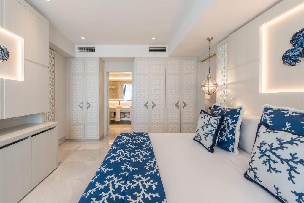 Mūsdienīgs guļamistabas interjers ar baltām sienām un zilo korālu rakstu spilveniem, radot elegantu un relaksējošu atmosfēru. Gaismas elementi un stūra skapis piešķir telpai papildu šarmu.
