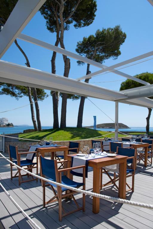 Restorāna terase ar koka galdiem un ziliem krēsliem, kas piedāvā skatu uz jūru un zaļo zāli. Saulains laiks un mierīga atmosfēra rada ideālu vidi atpūtai.