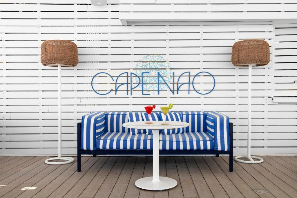 Mūsdienīga atpūtas zona ar zilu un baltu svītrainu dīvānu, baltu galdu un diviem kokteiļiem uz tā. Fons ir balts dēļu siena ar dekoratīvu uzrakstu "CAPENAO".
