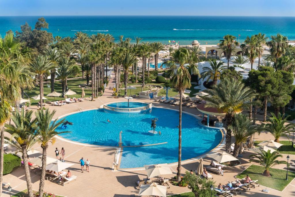 Vasaras kūrorta ainava ar palmām un peldbaseinu, kas atrodas blakus pludmalei un jūrai. Relaxējoša atmosfēra ar viesiem, kas atpūšas pie baseina un saulē.