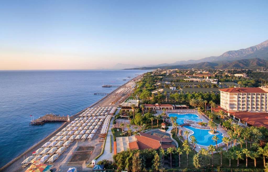 Pārskats uz pludmali ar saulrietu, kur redzams viesnīcas komplekss, baseins un pludmales gultas. Zilais jūras ūdens un kalnu fons piešķir gleznainu atmosfēru. panoramic view with beach and pools