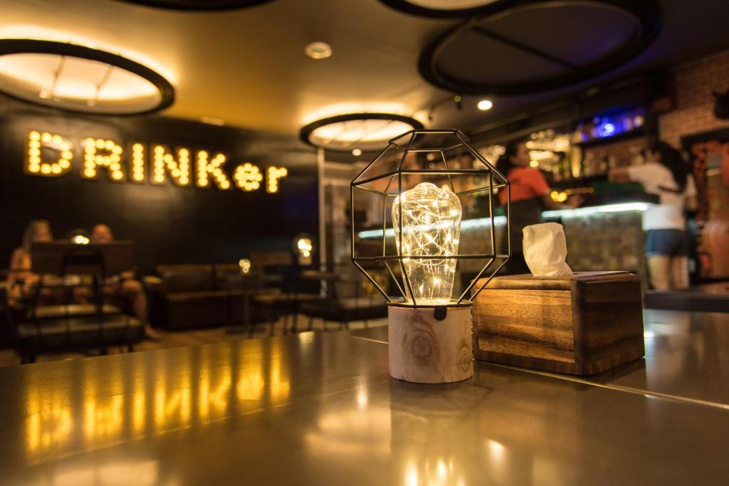 Kafejnīcas interjers ar stilīgu gaismekli un koka salvetu turētāju, fonā redzama dzīvīga atmosfēra ar cilvēkiem un izgaismotiem vārdiem 