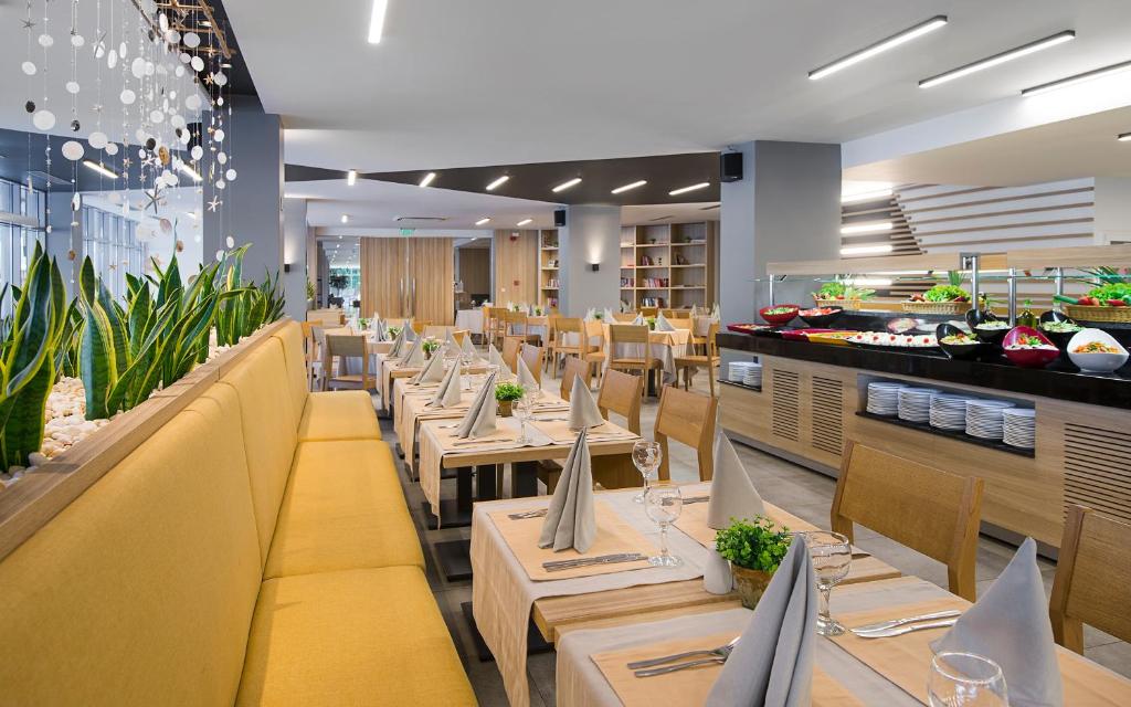 Mūsdienīgs restorāns ar gaišu interjeru, dzeltenām sēdvietām un zaļiem augiem, kas rada patīkamu atmosfēru. Uzkodu bārs ar daudzveidīgiem ēdieniem un dekoratīvām galda iestatījumiem.