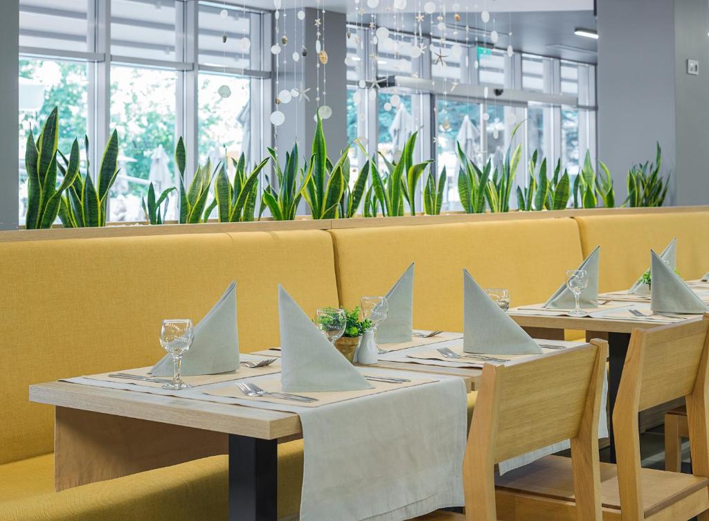 Mūsdienīgs restorāns ar dzeltenām mīkstajām mēbelēm un eleganti klātiem galdiem, kas rotāti ar zaļām augiem. Gaisīgs interjers ar lieliem logiem un modernu dekoru.