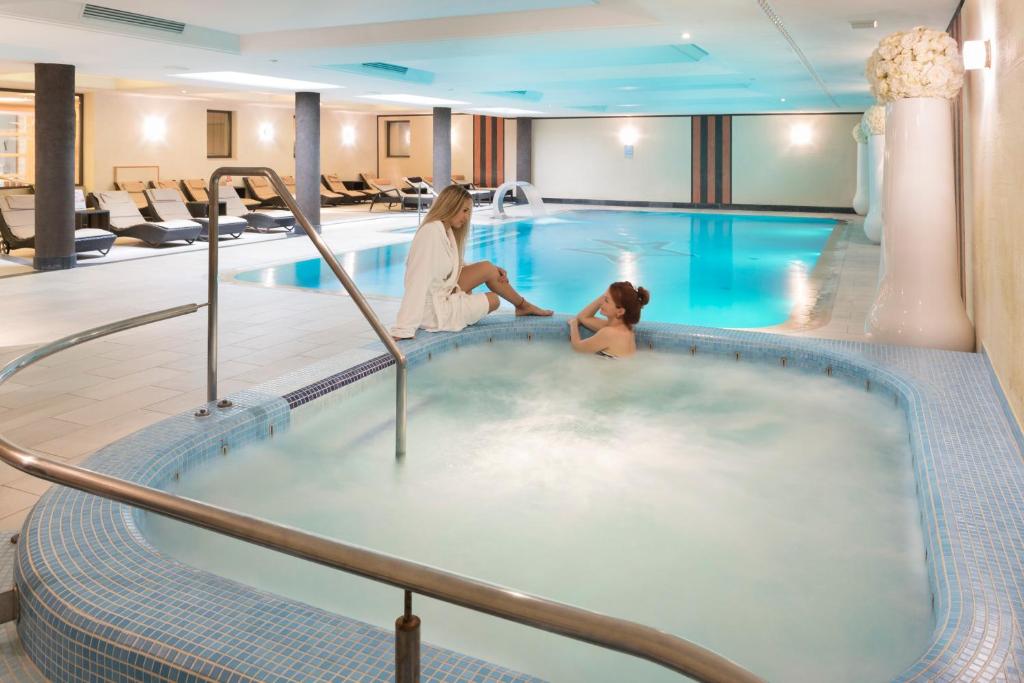 Spa telpa ar atpūtas baseinu un burbuļvannu, kur divas sievietes izbauda relaksāciju. Interjers ir mūsdienīgs ar komfortablām atpūtas vietām un patīkamu apgaismojumu.