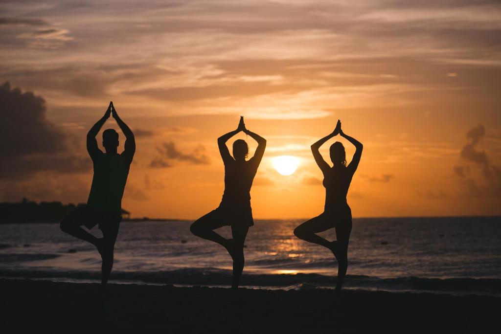 Trīs cilvēki veic jogas pozas pludmalē, pretī saulrietam. Silueti izceļas uz krāšņo debesu fona.