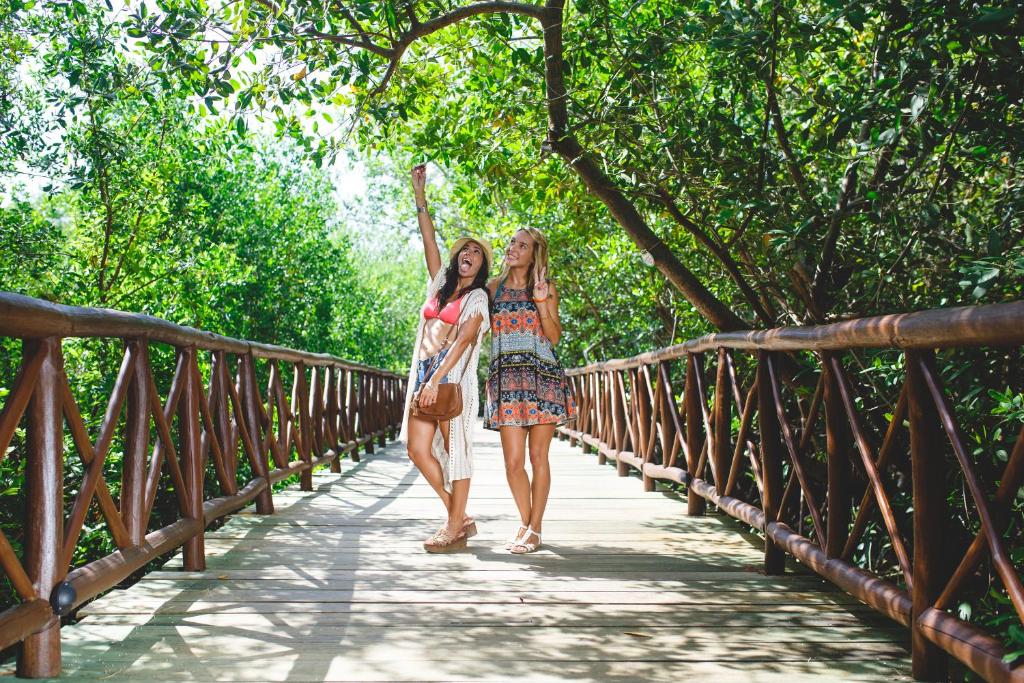 Divas draudzenes pozē uz koka tilta starp zaļām kokiem, izskatoties laimīgas un jautras. Viņas valkā vasaras apģērbus un izsaka pozitīvas emocijas.